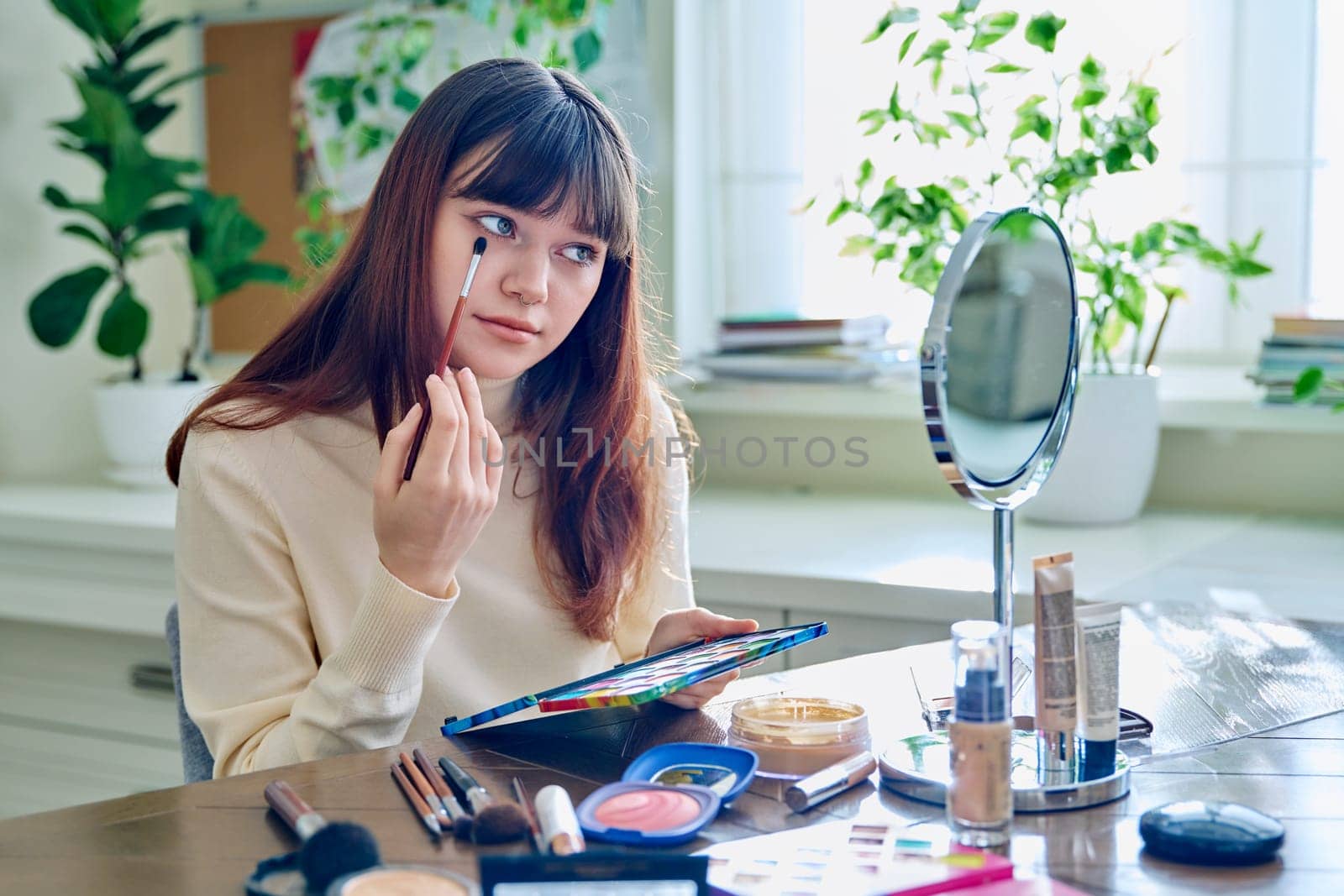 Young woman with cosmetic products on table, doing makeup looking in mirror at home. Cosmetics, fashion, youth, lifestyle, beauty concept