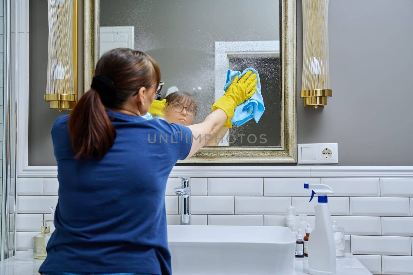 Woman cleaning bathroom, washing mirror with spray bottle and professional washcloth for cleaning glass without streaks. Housekeeping housework housecleaning cleaning service concept