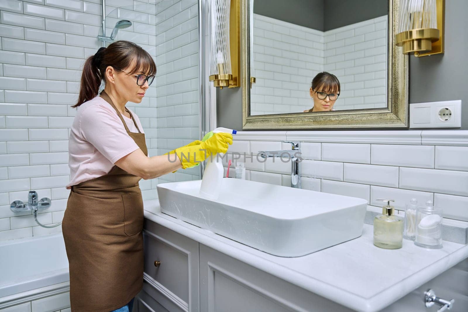 Woman in apron with detergent washcloth cleaning in bathroom, washing sink washbasin. Housewife cleaning house, cleaner service worker at workplace. Home hygiene housecleaning housekeeping housework