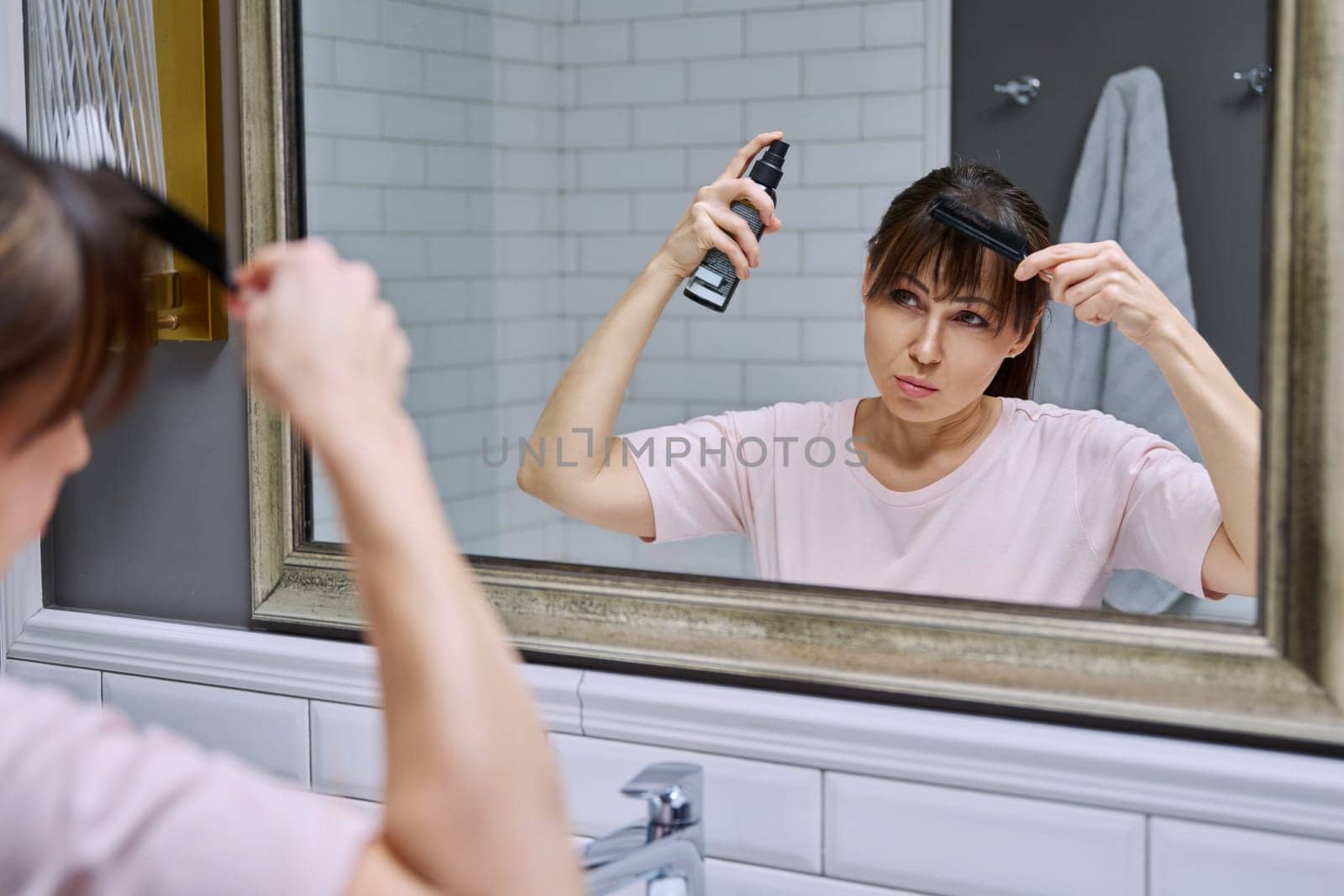 Middle-aged woman with setting spray for styling hair fixation comb looking in mirror, doing hair styling, in bathroom. Beauty, age, cosmetics, hair accessories