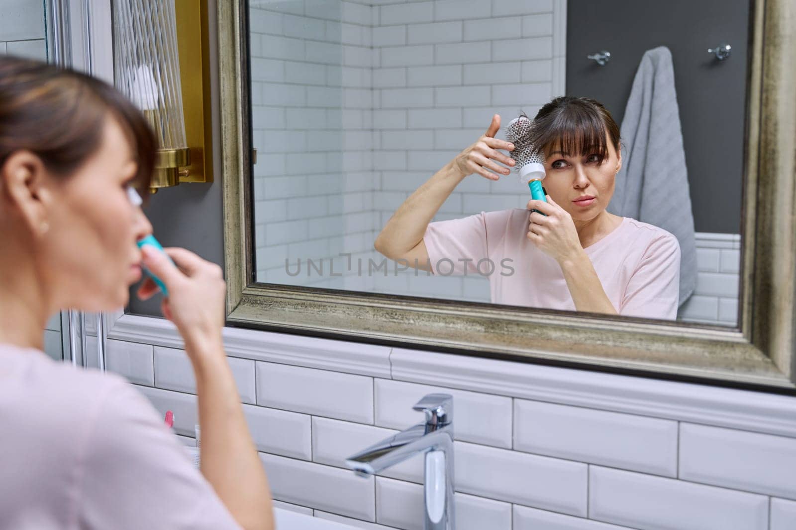 Middle-aged woman with comb looking in mirror, doing hair styling, in bathroom. Beauty, age, cosmetics, hair accessories
