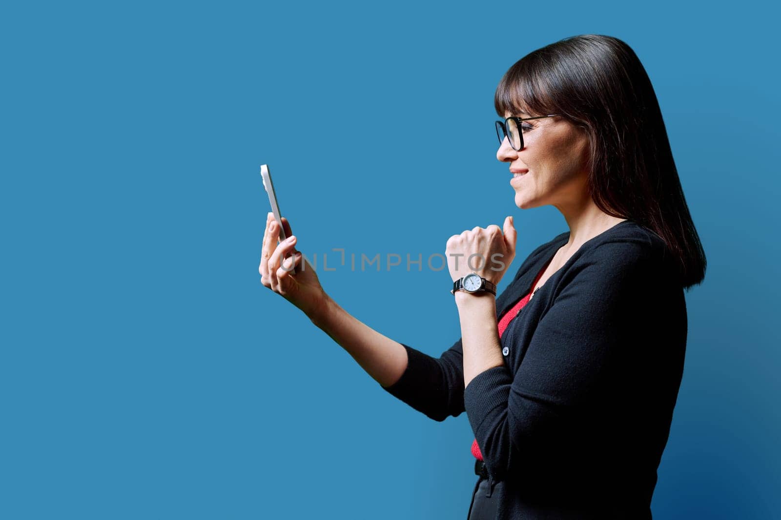 Profile view middle aged smiling woman using smartphone on blue background. Mature female looking at phone in hands. Technologies mobile apps applications internet work business leisure communication