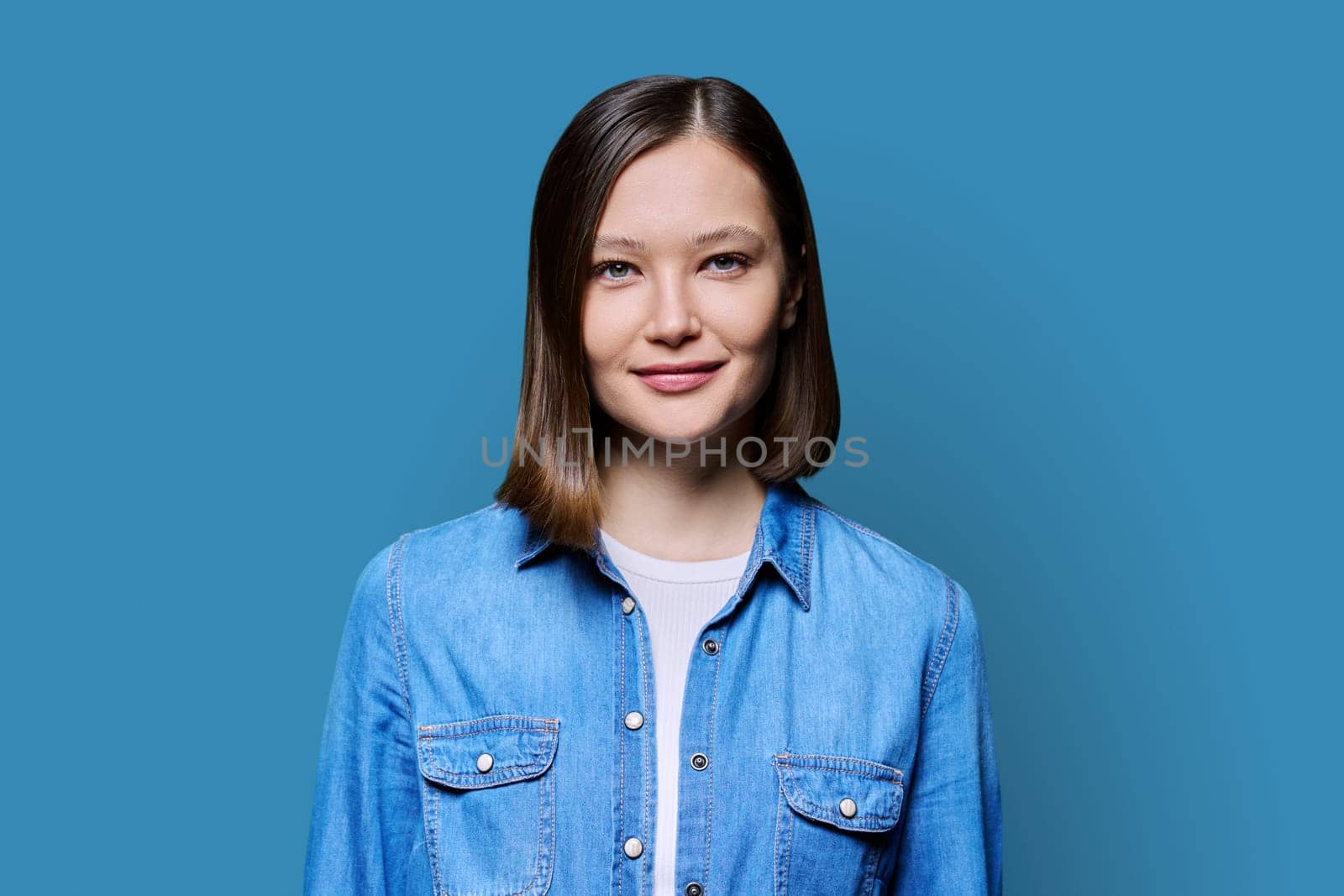 Young beautiful cheerful woman on blue studio background. Smiling attractive 20s female in denim looking at camera. Lifestyle, fashion beauty style, youth business work services, education, healthcare