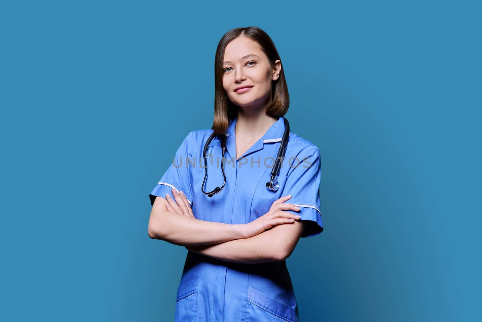 Portrait of young confident smiling female nurse with stethoscope, crossed arms, looking at camera on blue studio background. Medical services, health, professional assistance, medical care concept