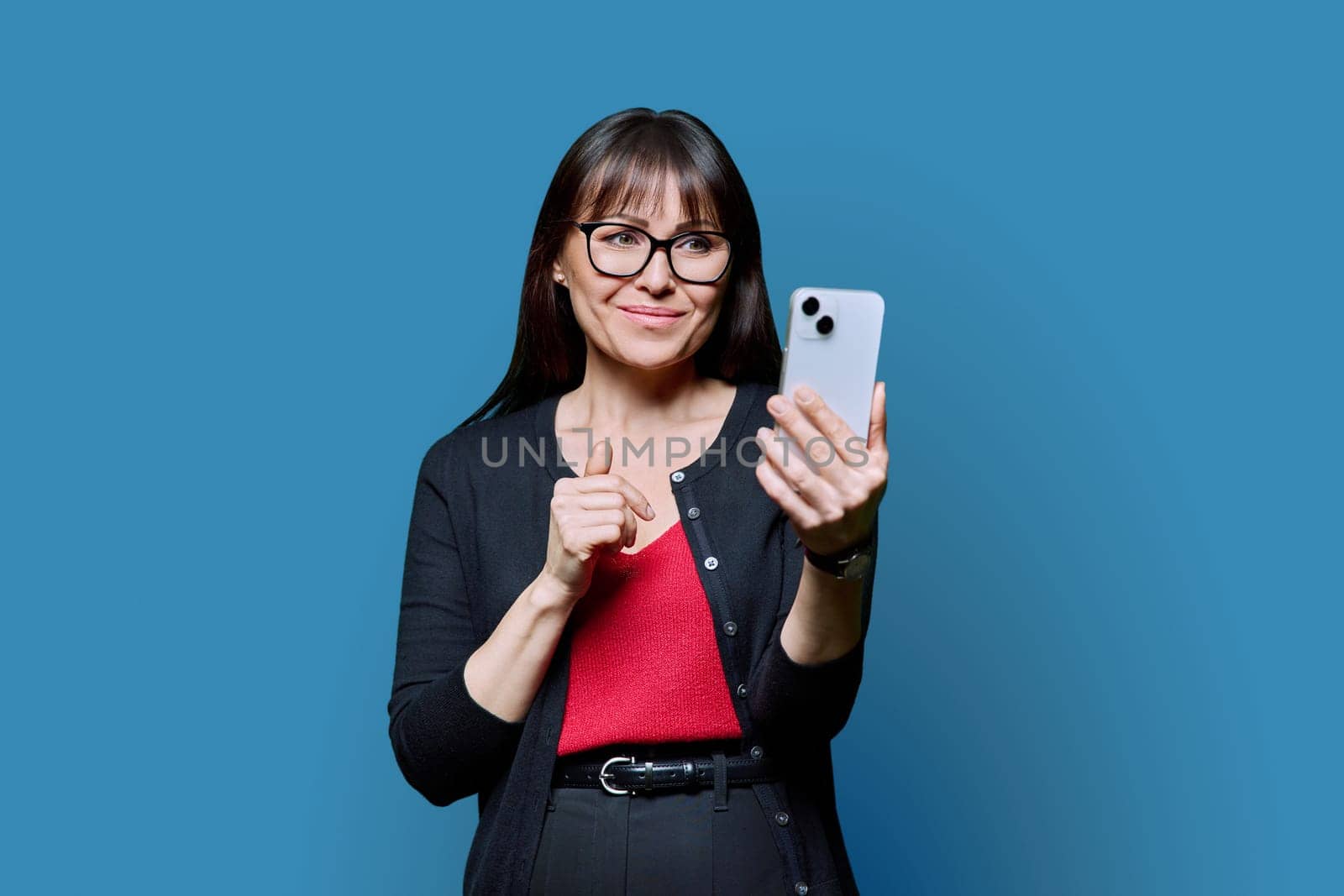 Middle aged smiling woman using smartphone on blue background. Mature female looking at screen holding phone in hands. Technologies mobile applications internet work business leisure communication