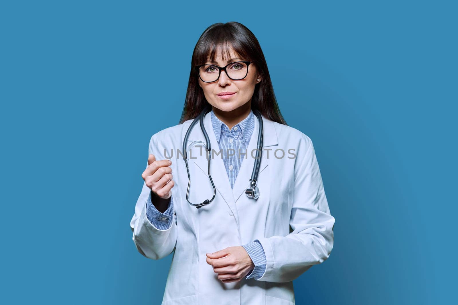 Confident friendly smiling middle-aged female doctor in white lab coat with stethoscope, looking at camera on blue studio background. Healthcare, medicine, staff, treatment, medical services concept