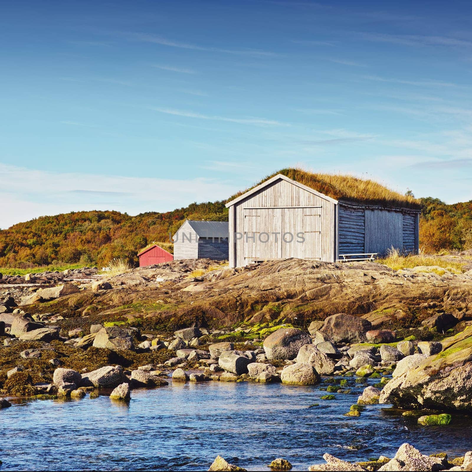 Norway, house and outdoor in field, lake and rocks for travel and holiday in background with hills. Shed, grass and blue sky for cottage, serenity and sunlight for nature in season for countryside by YuriArcurs