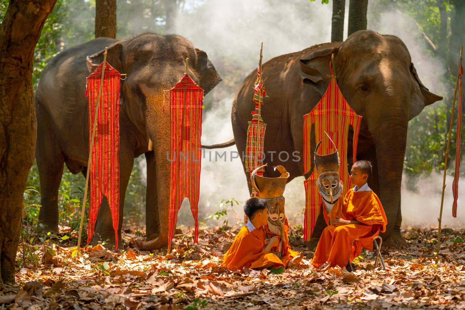 Two boys with Pee Ta Khon dress hold mask and talk together in front of elephant and traditional flags on background in forest.