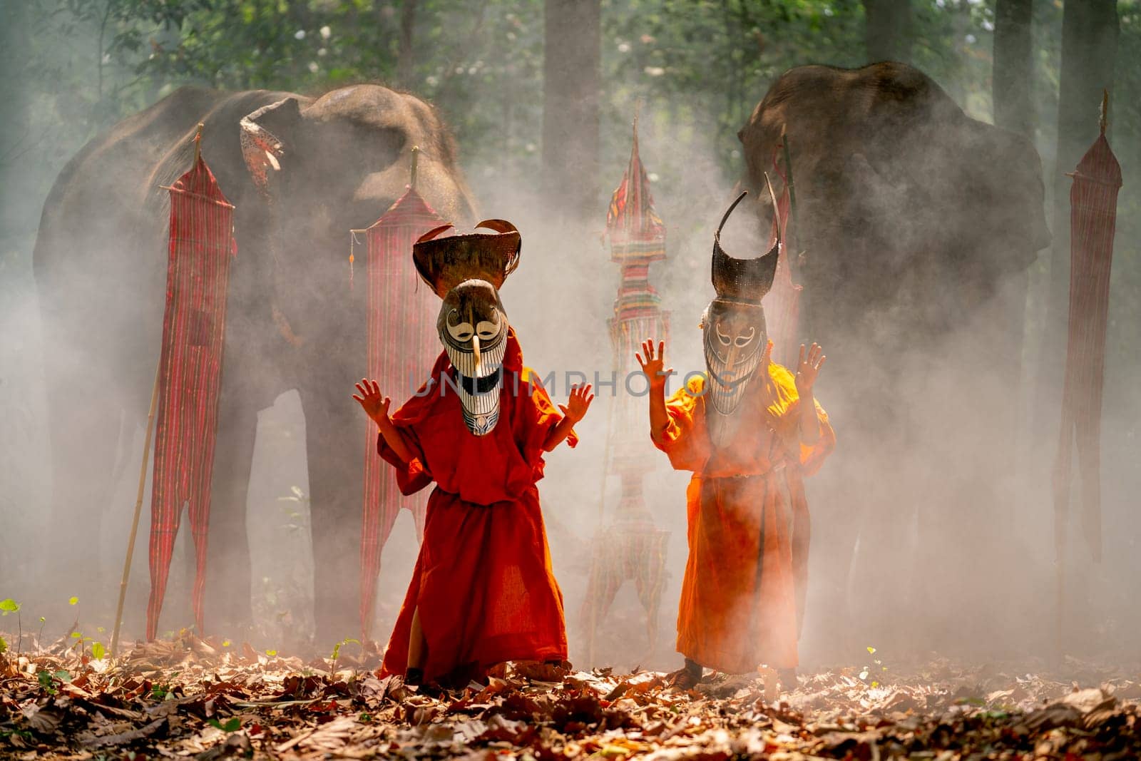 Two boy wear Pee Ta Khon, traditional culture art as ghost of Asian culture, dress and look at camera and elephants stand in the background and they stay in forest. by nrradmin