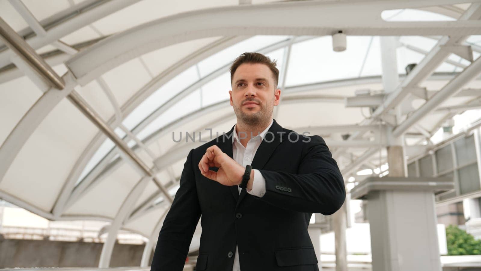 Skilled business man looking watch while walking to train station for going to workplace at bridge. Caucasian manager checking time for important appointment and walk along modern urban city. Urbane.