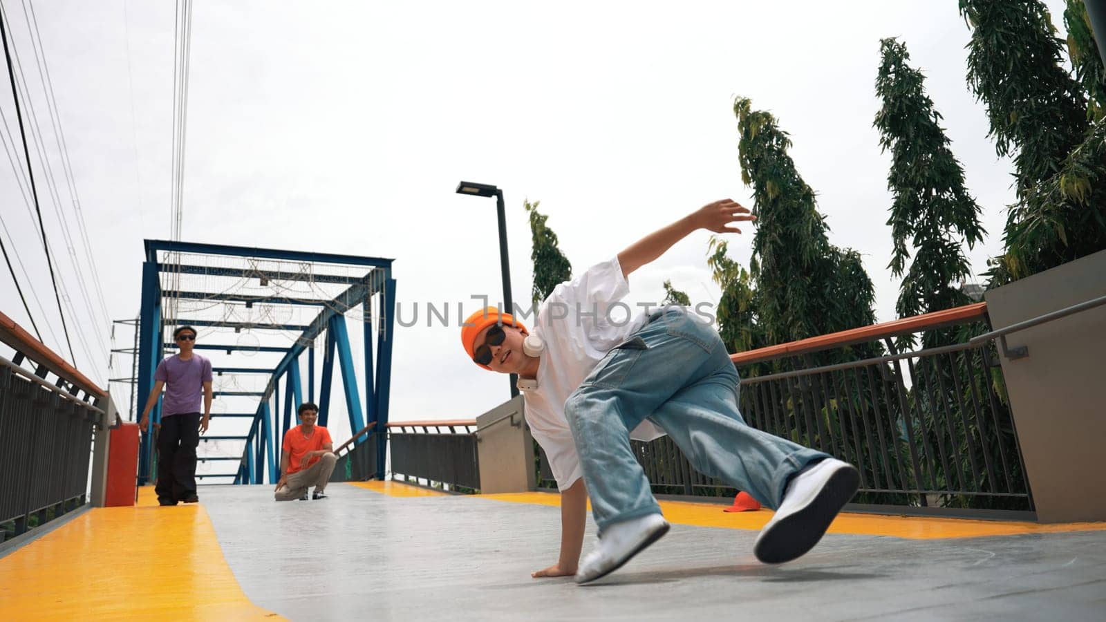 Panorama shot of professional hipster perform b-boy performance and energetic footstep. Skilled happy man practice break dance while friend cheering him at bridge. Outdoor sport 2024. Sprightly.