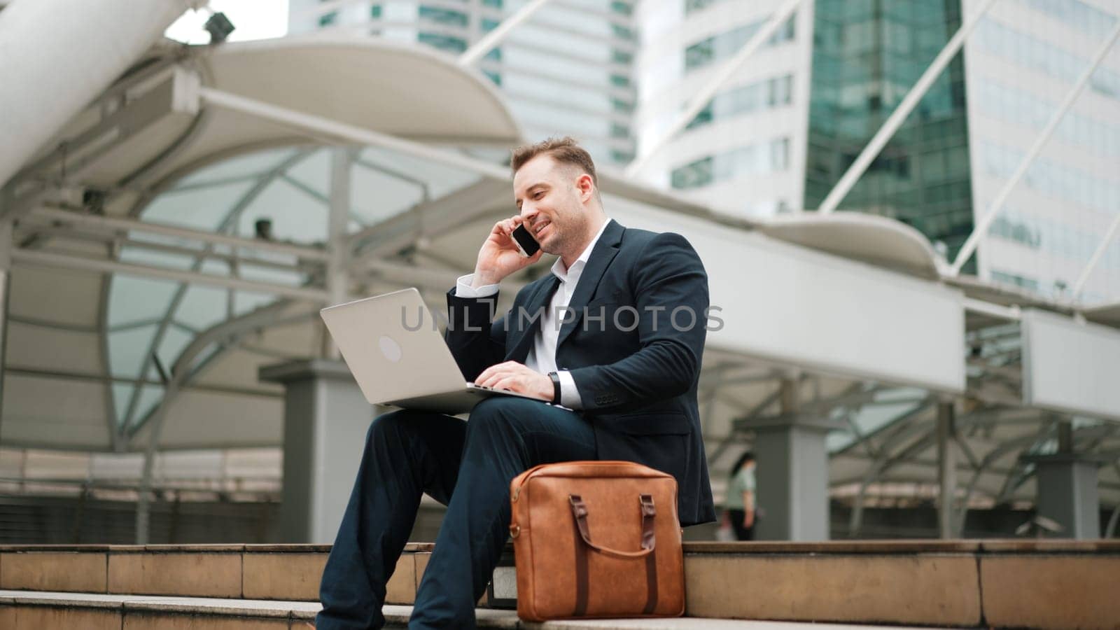 Caucasian business man sitting at stairs while using laptop and phone. Urbane. by biancoblue