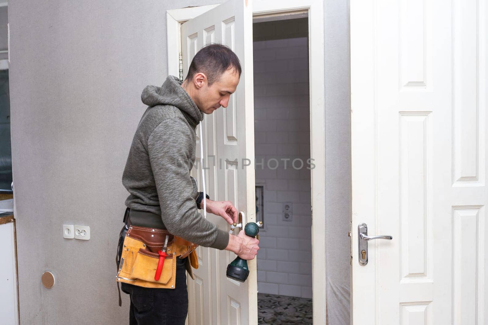Maintenance Concept. Focused serious young locksmith with tattoo on hand wearing blue uniform standing on one knee in doorway installing doorknob handle, repairing wooden front door entrance. High quality photo