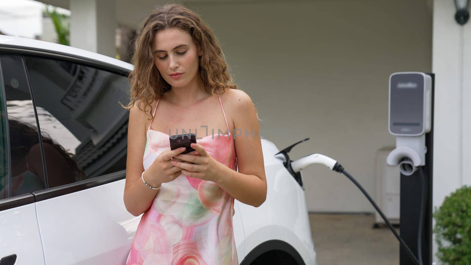 Modern eco-friendly woman recharging electric vehicle from home EV charging station. Innovative EV technology utilization for tracking energy usage to optimize battery charging at home. Synchronos