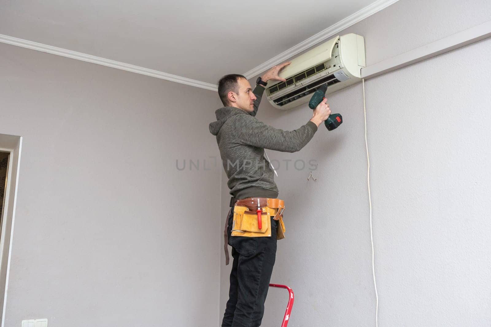 Professional electrician man with screwdriver maintaining, cleaning modern air conditioner indoors. Young technician standing on a ladder in the room repairing or installing air conditioner. High quality photo