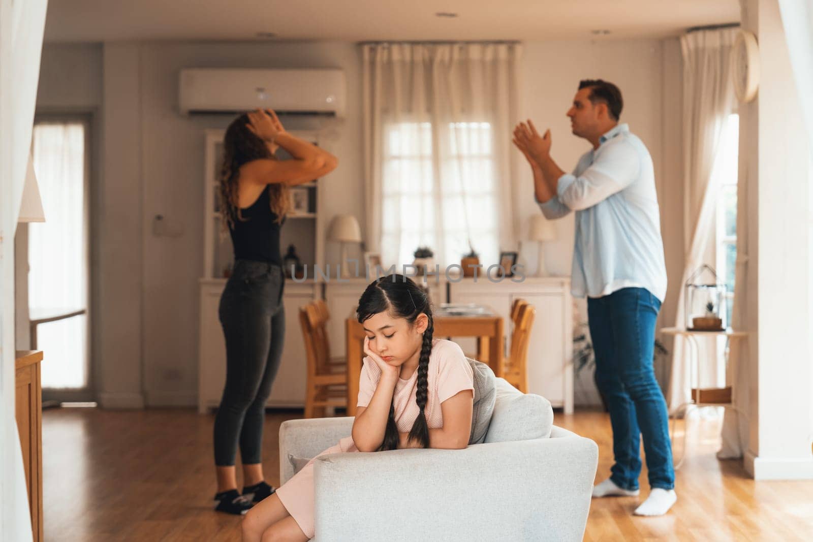 Annoyed and unhappy young girl sitting on sofa trapped in middle of tension by her parent argument in living room. Unhealthy domestic lifestyle and traumatic childhood develop to depression.Synchronos