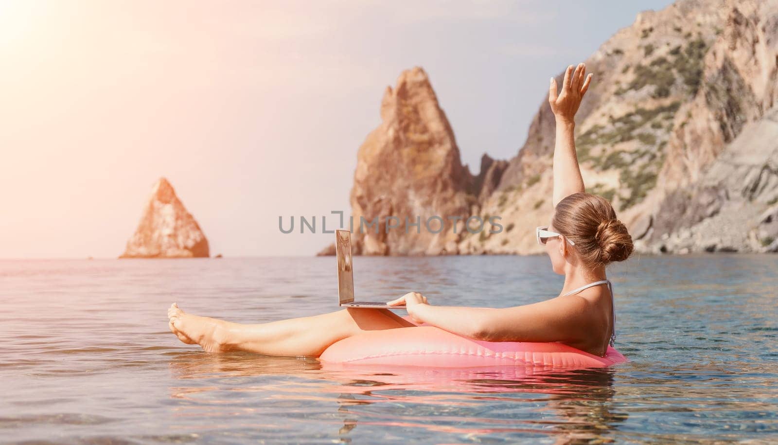 Woman freelancer works on laptop swimming in sea on pink inflatable ring. Pretty lady typing on computer while floating in the sea on inflatable donut at sunset. Freelance, remote work on vacation