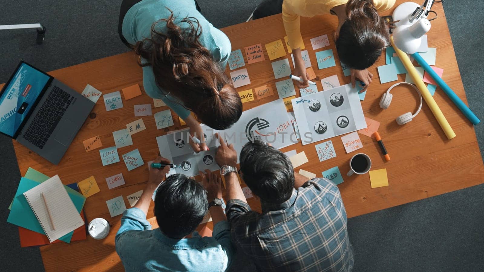 Top down aerial view of skilled designer hold logo while discuss with team. Aerial business group working together sharing idea about logo graphic design by using sticky notes and tablet. Symposium.