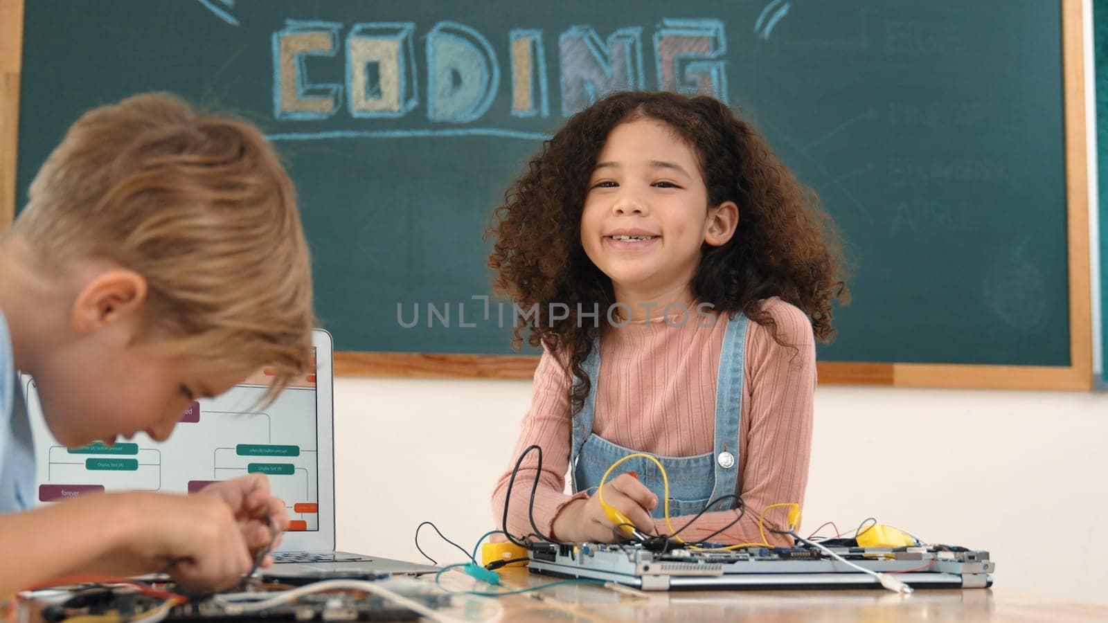 Girl standing while fixing electronic board by using screwdriver. American student and happy caucasian boy working together to inspect electric system. Curious children working on board. Pedagogy.