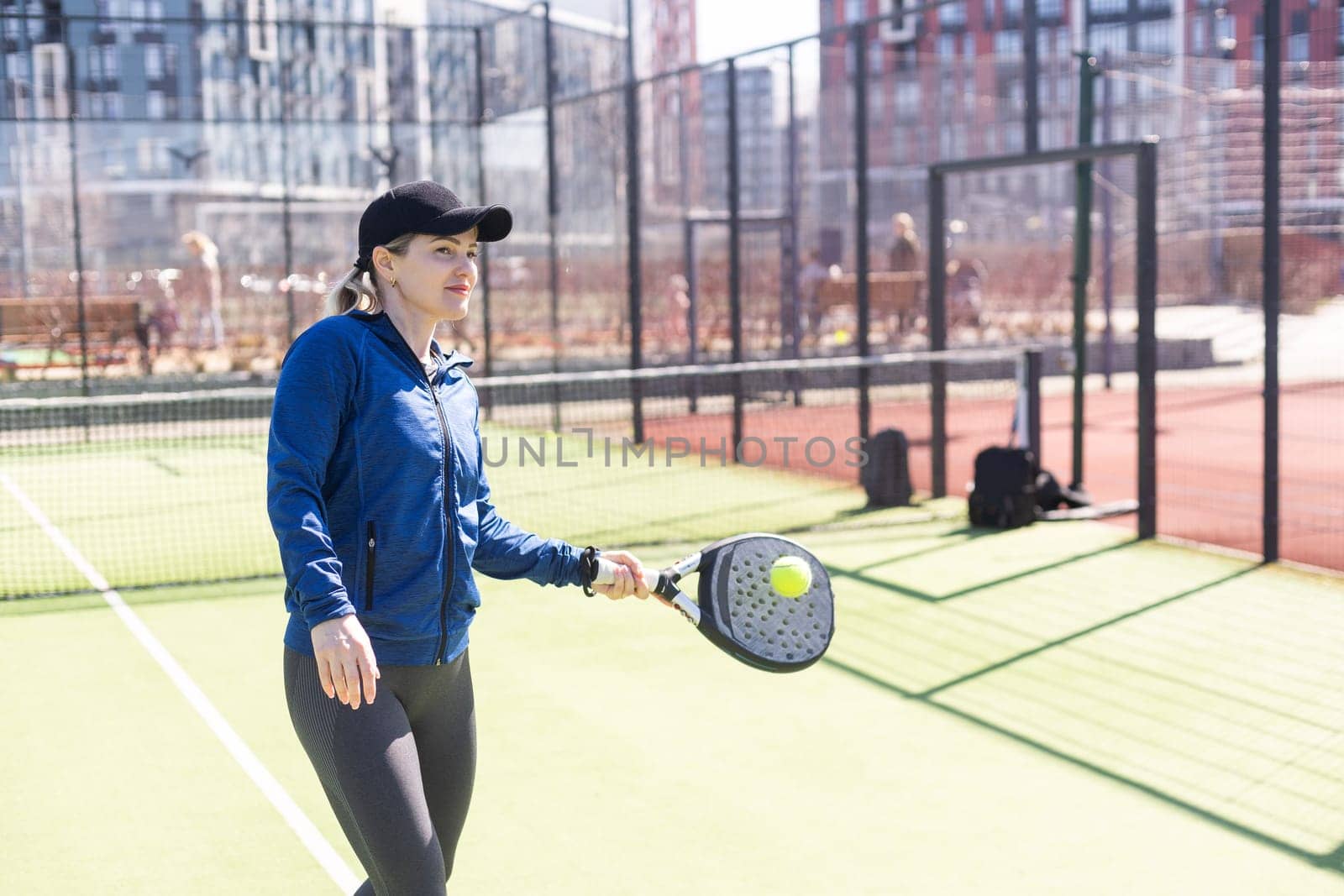 A girl in sportswear is training on a paddle tennis court. The girl is hitting the ball against the glass to make a rebound. Concept of women playing paddle. by Andelov13