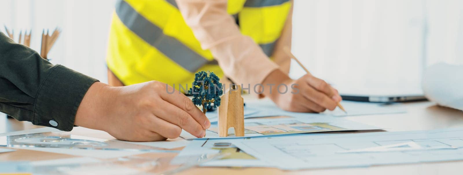 Professional architect and engineer collaborate to design eco-friendly house on meeting table with blueprint, wooden house block and tree model scatter around. Closeup. Delineation.