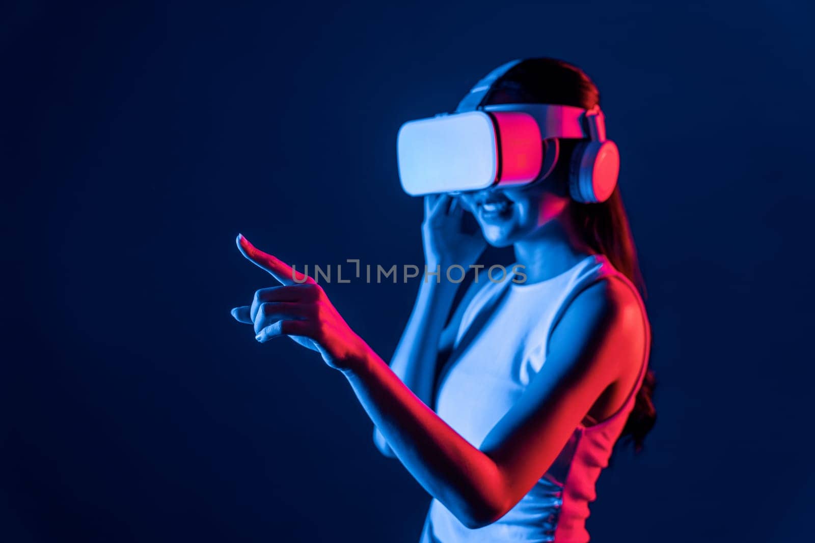 Smart female stand with surrounded by cyberpunk neon light wear VR headset connecting metaverse, futuristic cyberspace community technology. Woman using finger pointing virtual object. Hallucination.