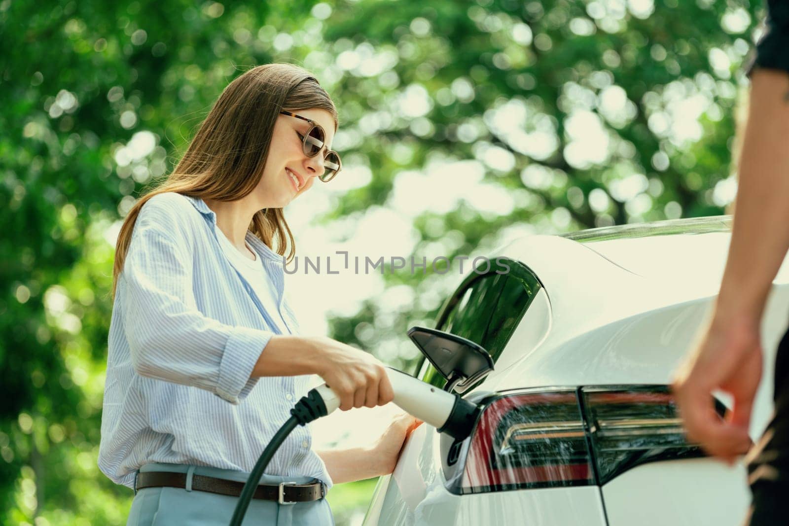 Lovely young couple wearing sun glasses recharging battery for electric car during road trip travel EV car in natural forest or national park. Eco friendly travel during vacation and holiday. Exalt