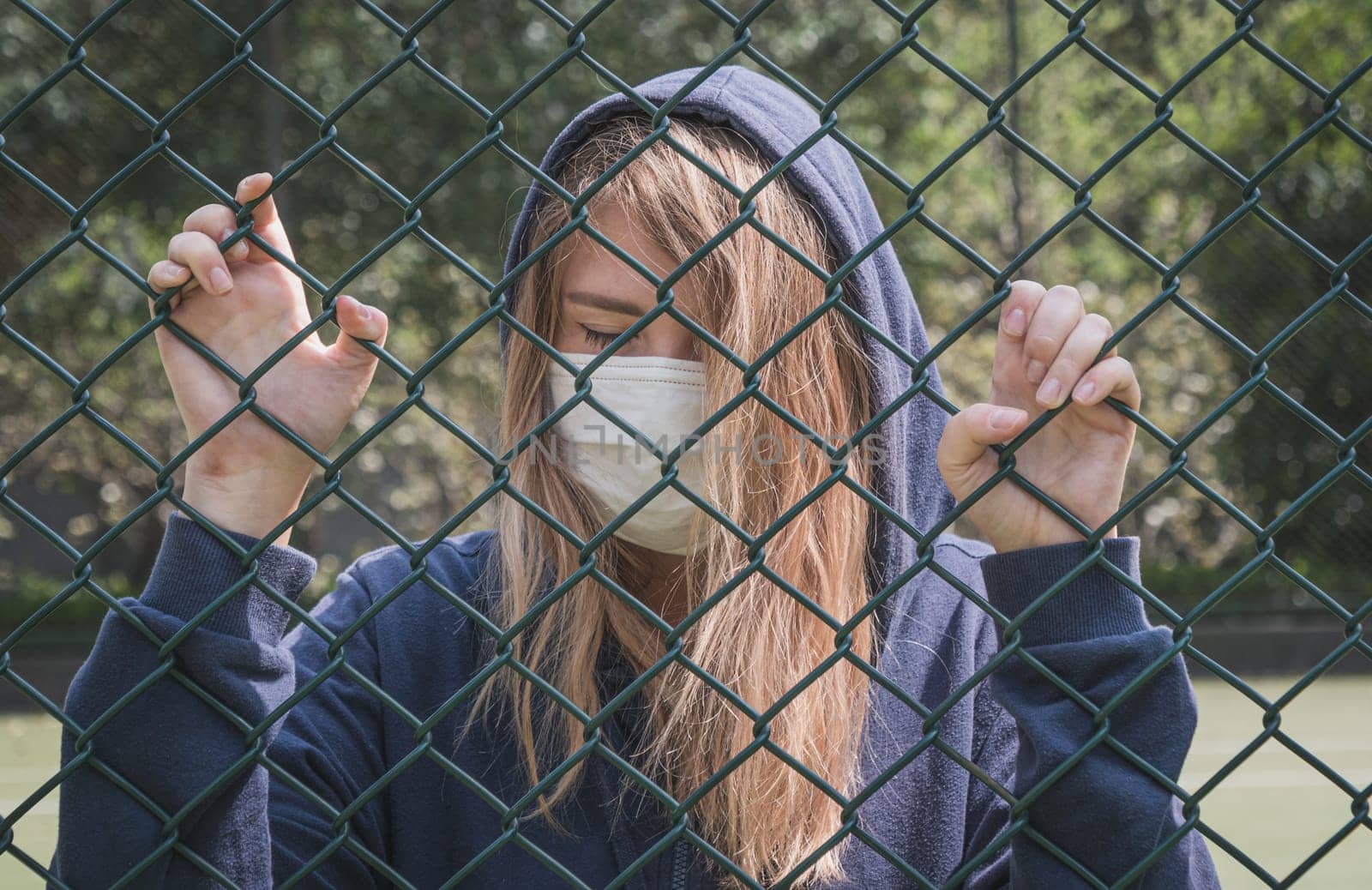 Close-up shot of young woman wearing Protective Face Mask stand behind fence border. COVID-19 coronavirus infection pandemic disease virus
