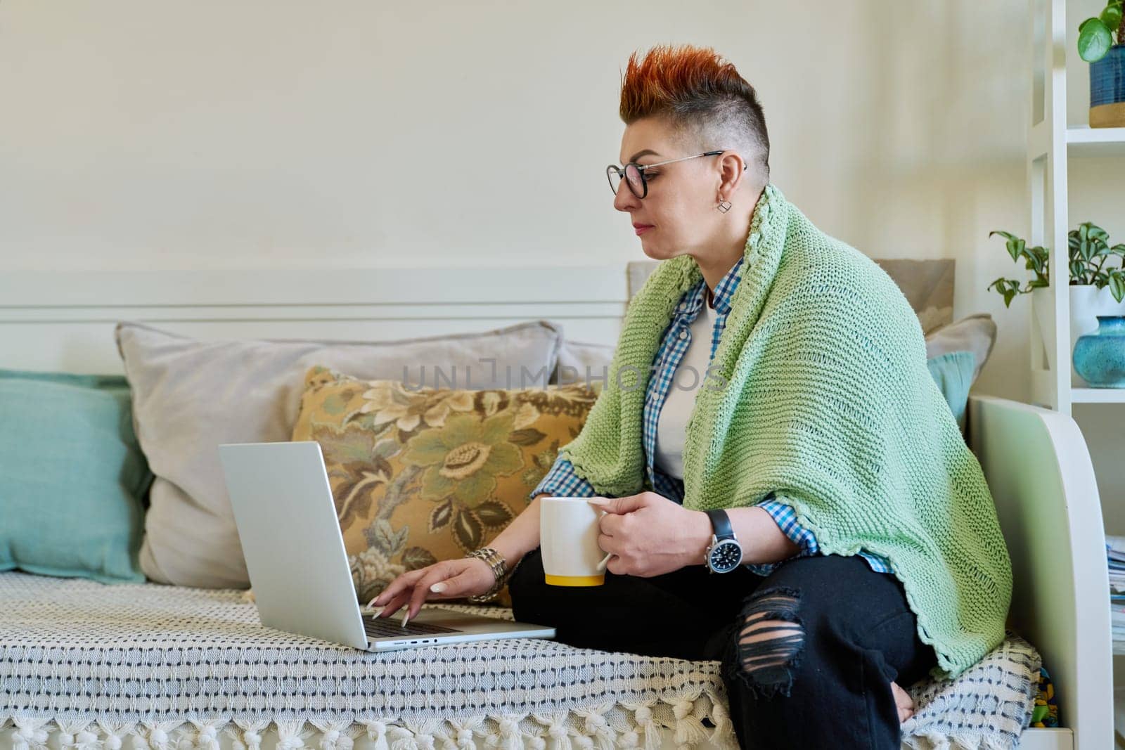 Mature woman with warm blanket holding cup of hot drink looking at laptop screen sitting on couch at home. Home coziness comfort, cold seasons, lifestyle, technology for leisure work communication