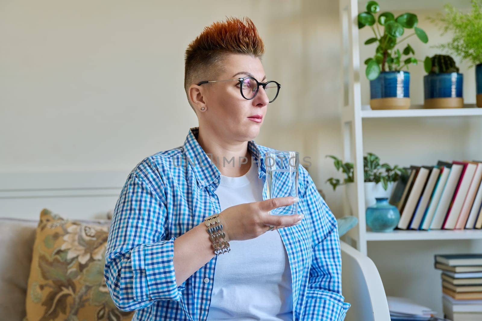 Middle aged woman with a glass of water sitting on the couch at home. Health, nutrition, beauty, 40s age concept