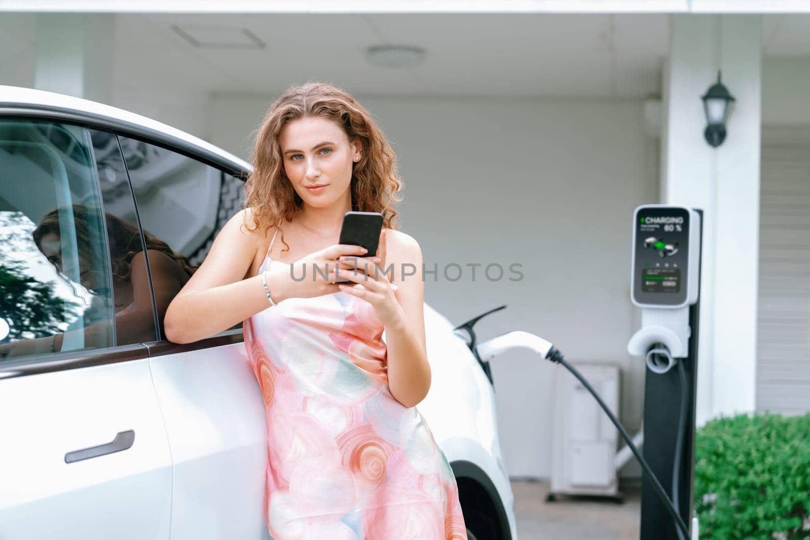 Modern eco woman recharge EV car at home with phone. Synchronos by biancoblue