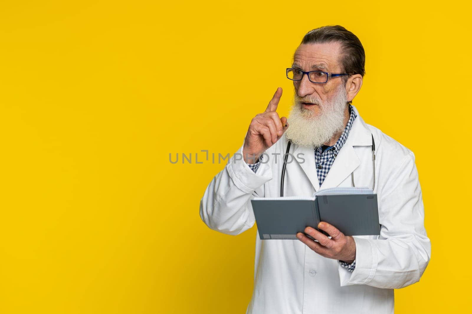 Good idea. Thoughtful senior mature doctor cardiologist man making notes, writing down thoughts with pen into notepad notebook diary, to do list. Apothecary pharmacy grandfather on yellow background