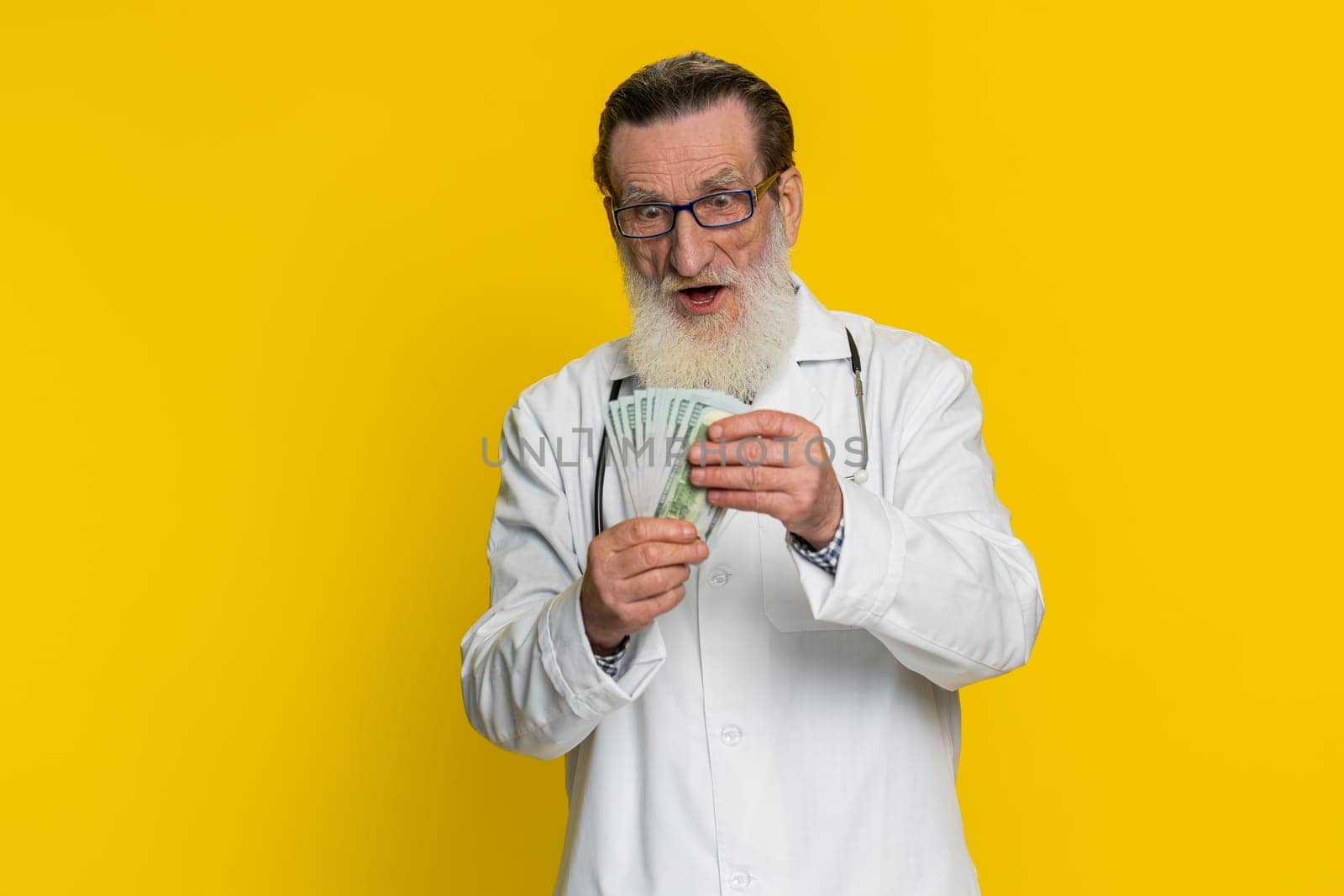 Rich happy senior doctor cardiologist man waving money dollar cash banknotes bills like a fan, success business career, big income wealth. Apothecary pharmacy grandfather isolated on yellow background