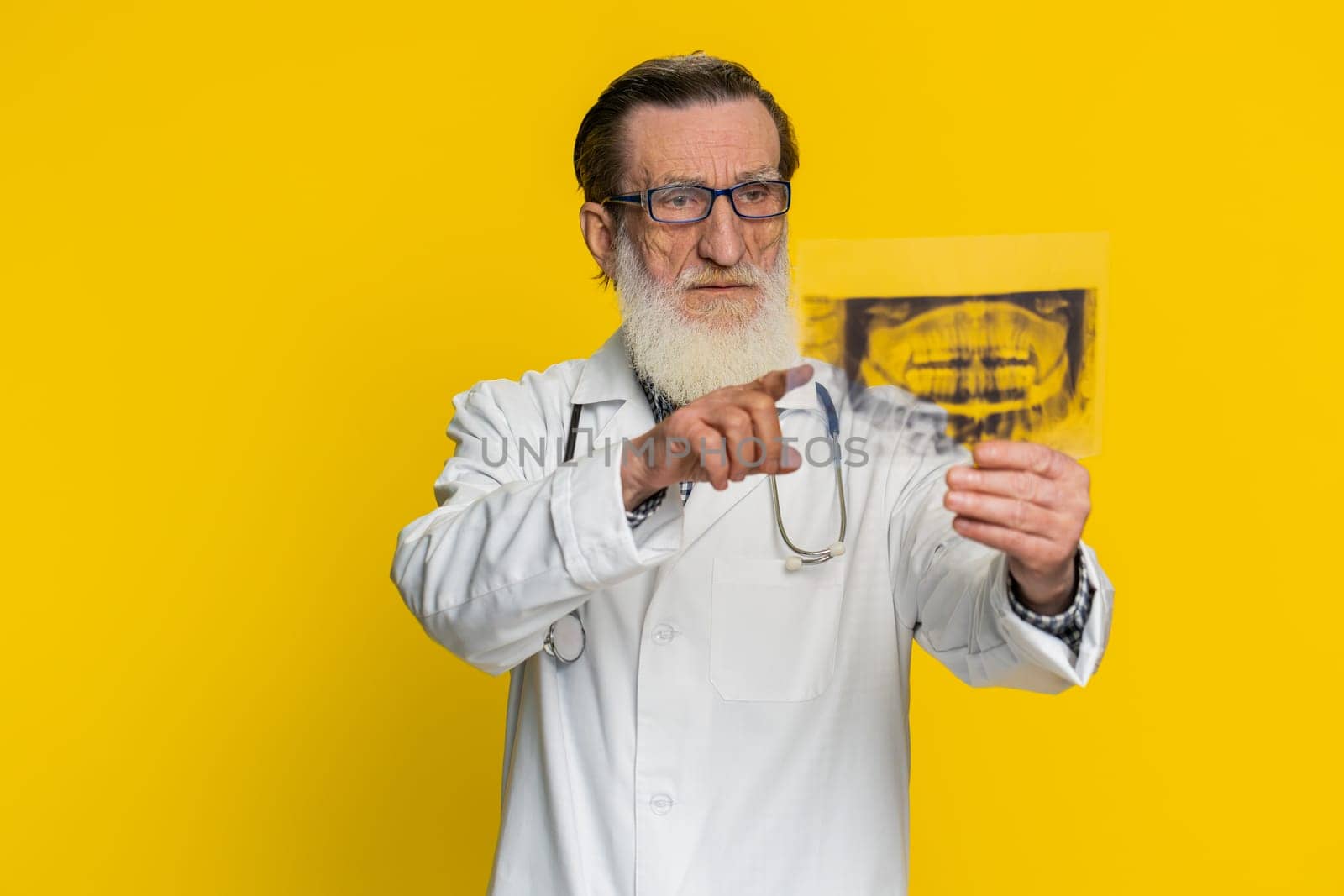 Senior elderly doctor orthodontist man examines a panoramic x-ray picture of the jaw teeth. 3D model of the patient's mouth, MRI scan. Dentistry oral care. Stomatology grandfather on yellow background