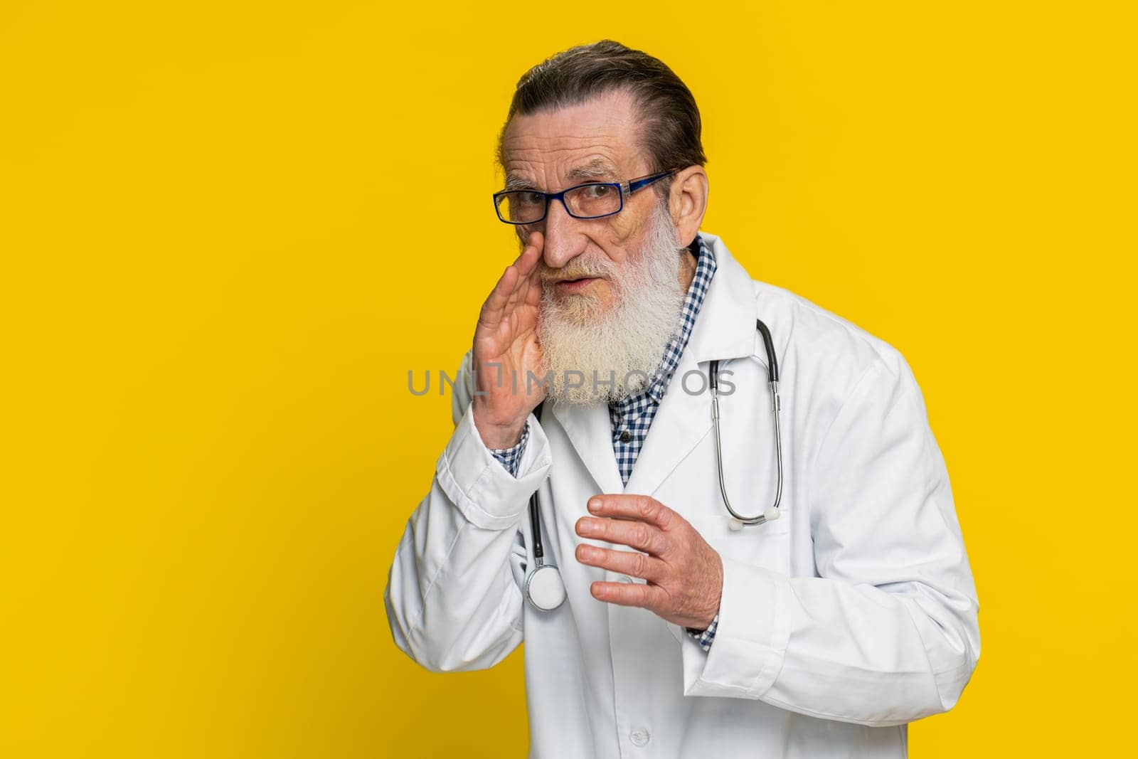 Keep my secrets, silence. Senior doctor cardiologist man whisper news rumors holding hands near mouth, share gossip quiet. Grandfather telling interesting confidential information on yellow background