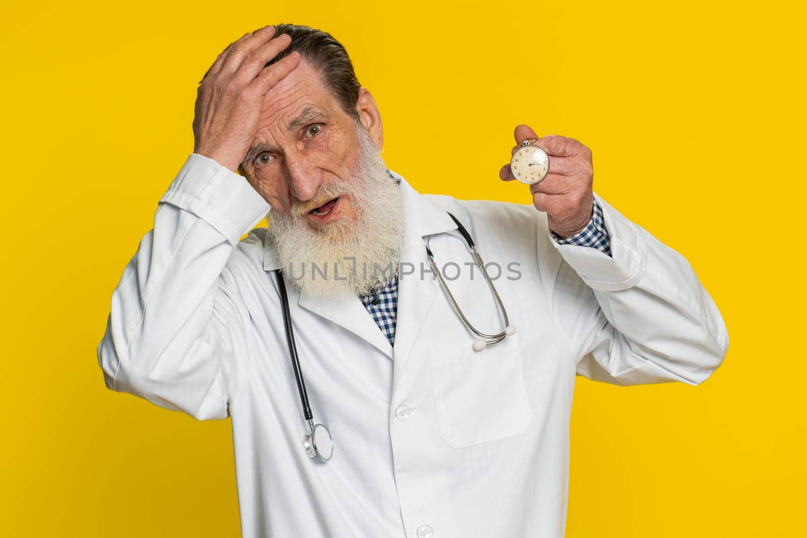 Mature doctor cardiologist man with anxiety checking time on pocket watch, running late to work being in delay deadline. Pharmacy old grandfather worrying to be punctual isolated on yellow background