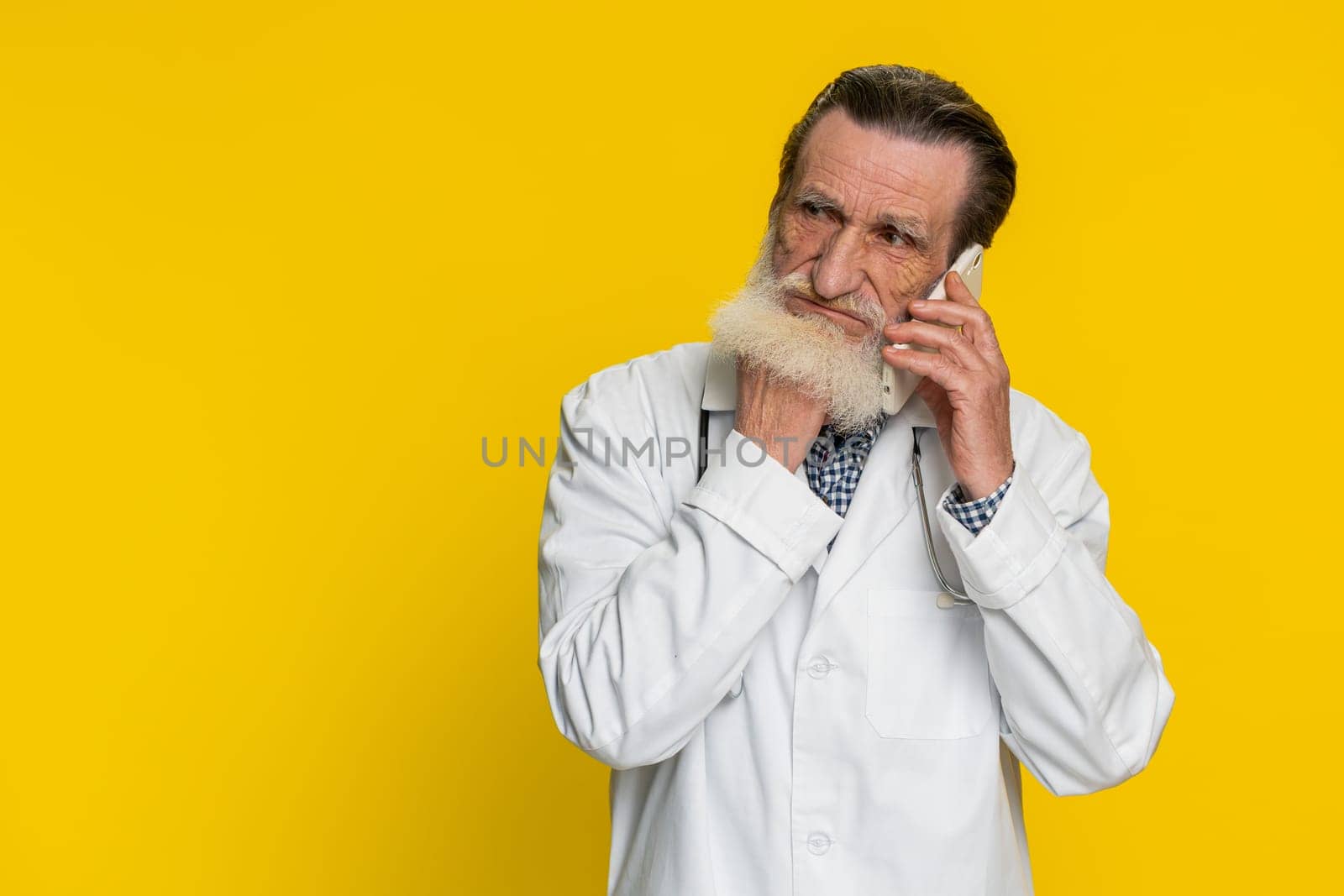 Disinterested senior doctor cardiologist man having annoyed boring talk on smartphone. Tired sleepy apothecary pharmacy grandfather freelancer talking on mobile phone with friend on yellow background