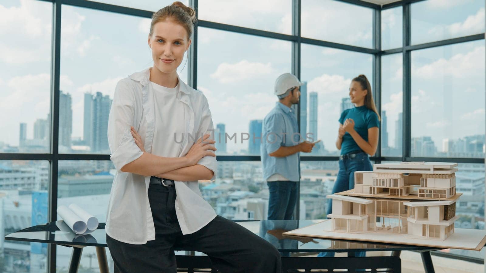 Young beautiful businesswoman sitting with arm folded while colleague discussion about house construction. Smart skilled architect engineer looking and smiling at camera with confident. Tracery