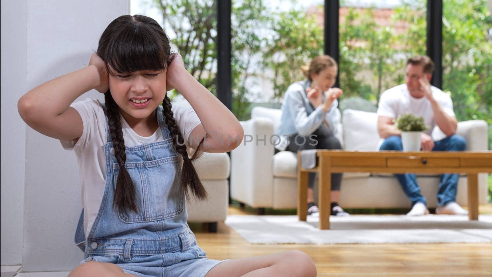 Stressed and unhappy young girl huddle in corner, cover her ears blocking sound of her parent arguing in background. Domestic violence at home and traumatic childhood develop to depression. Synchronos