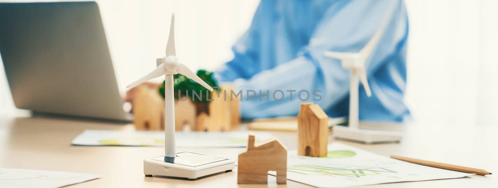 Windmill model represented using clean energy and wooden block represented eco house was scatter around on the table in front of businessman working on laptop. Blurring background. Delineation.