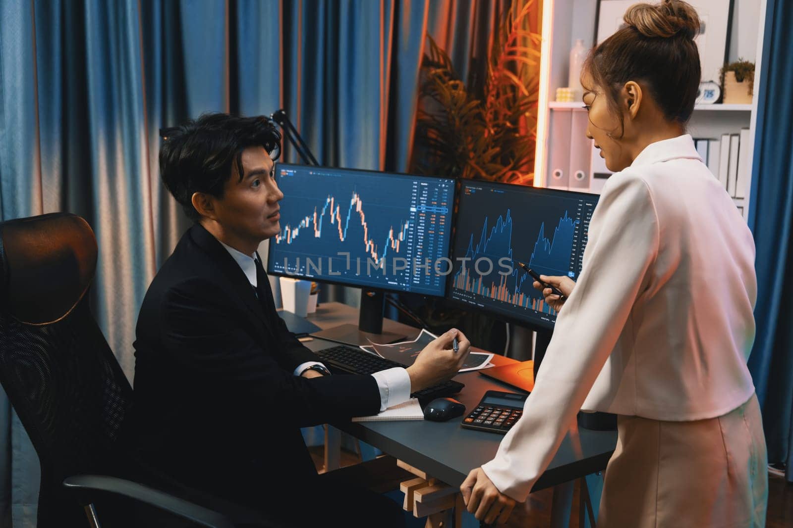 Asian businesswoman standing to discuss with dynamic stock exchange graph with partner to taking note currency rate on profit value online website program application at neon light at night. Infobahn.