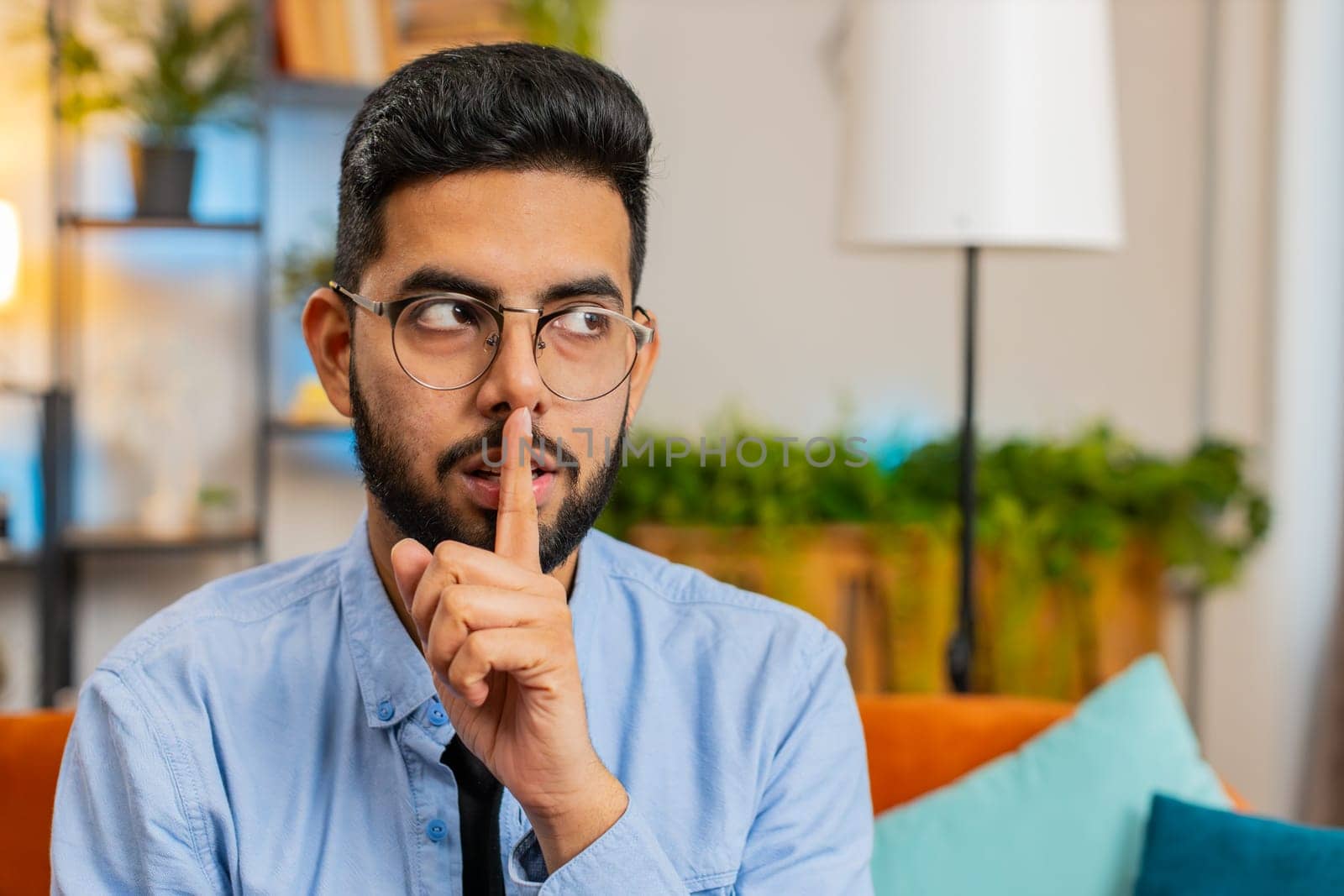 Shh be quiet please. Portrait of young Indian man presses index finger to lips makes silence gesture sign do not tells secret, stop talk gossip, confidential privacy. Guy sitting on couch at home room
