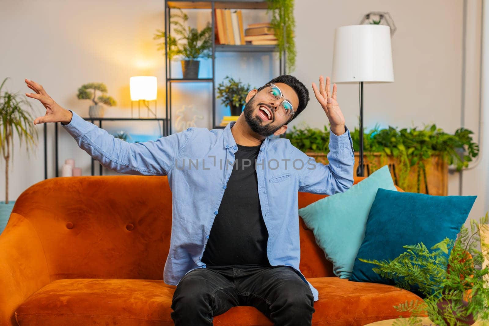 Energetic young Hispanic man singing and dancing with hands sitting on couch in living room. Carefree Indian guy enjoying favorite music and having fun while staying alone at home during weekend.