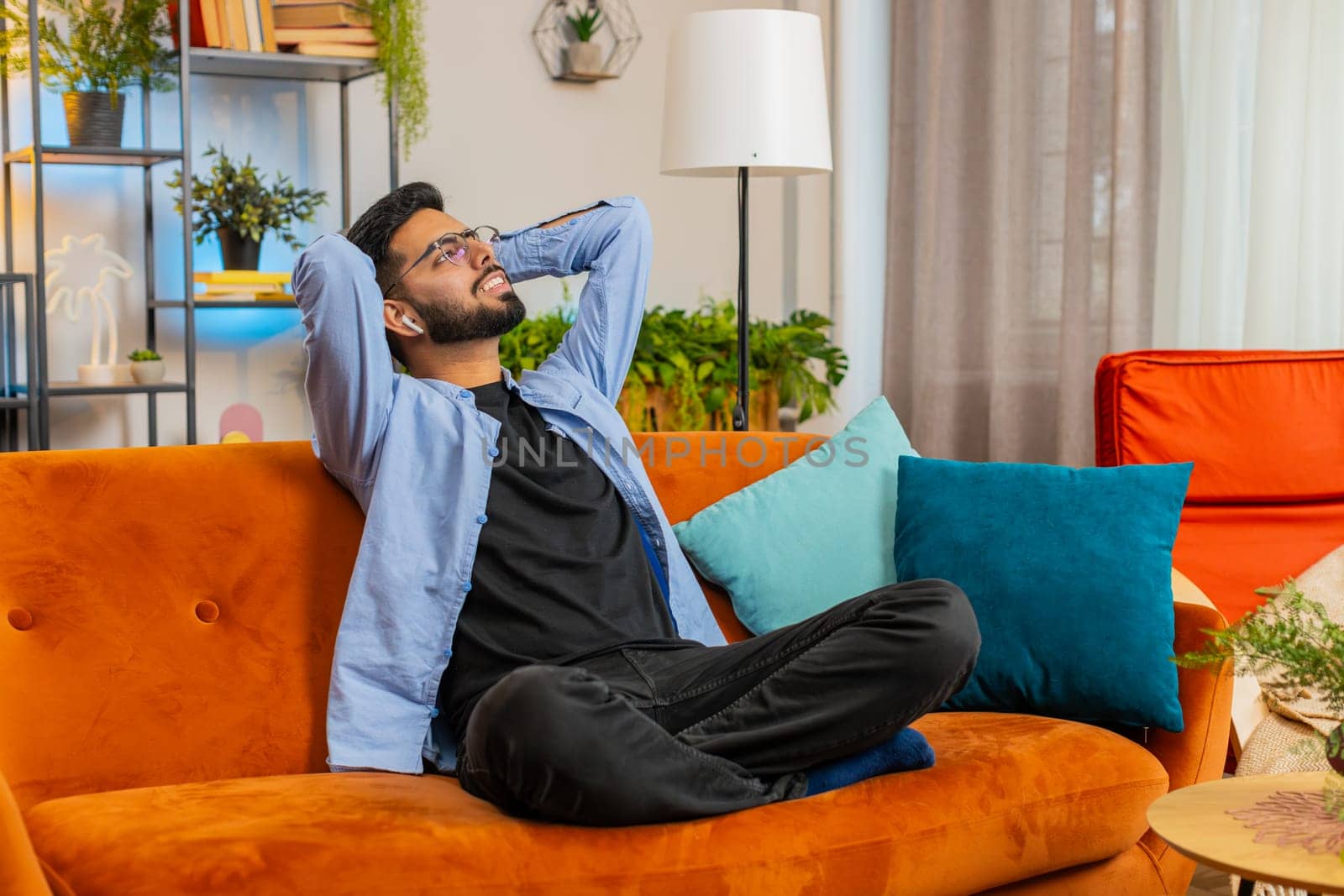 Happy relaxed young Indian man resting on couch with wireless earphones listening to ambient calming tranquil music from online playlist. Smiling Arabian guy resting with hands behind head in room.