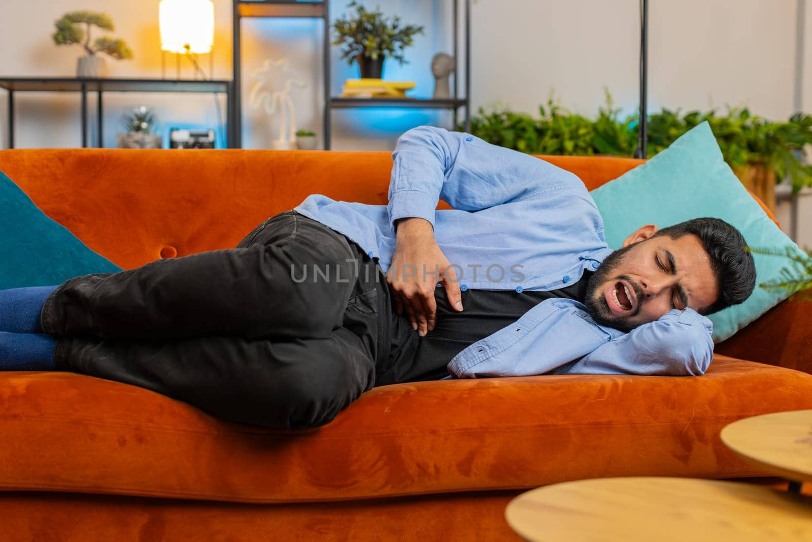 Indian young man lying on couch feeling sudden strong abdominal stomach ache, gastritis problem. Upset Arabian guy having symptom poisoning diarrhea indigestion peptic ulcer pancreatitis at home