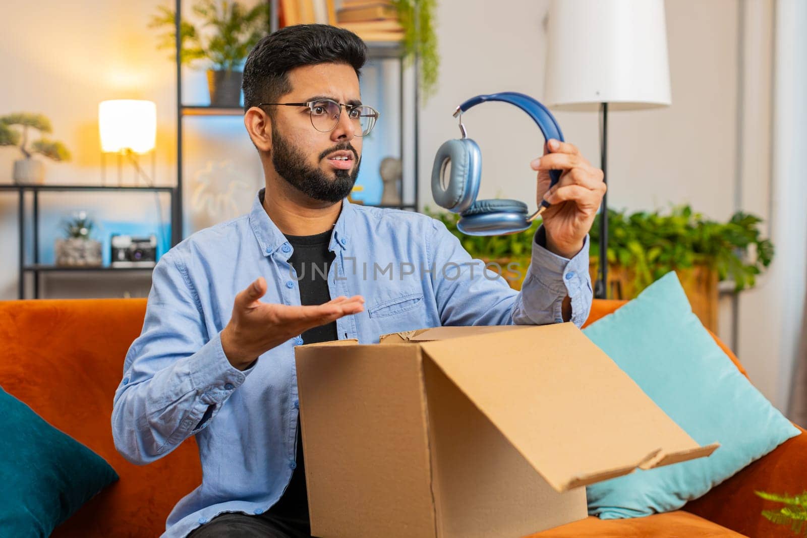 Dissatisfied annoyed Indian male customer open cardboard box receive damaged broken wireless headphones, shocked young man consumer having problem complaint frustrated with bad order delivery at home.