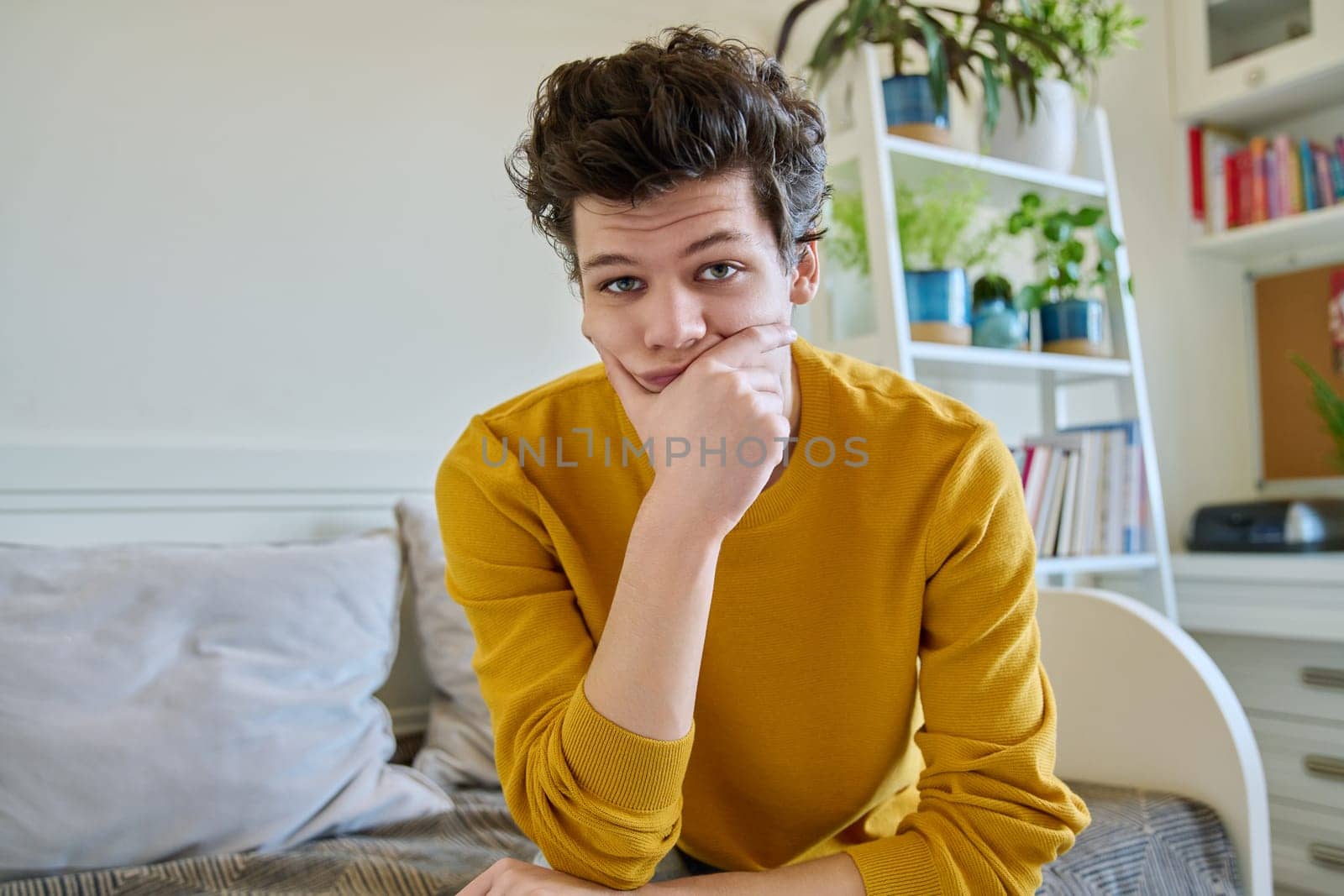 Portrait of handsome smiling guy looking at camera, web cam view, sitting on couch at home. Young man in yellow with curly hair, university college student. Lifestyle, youth 19-20 years old concept