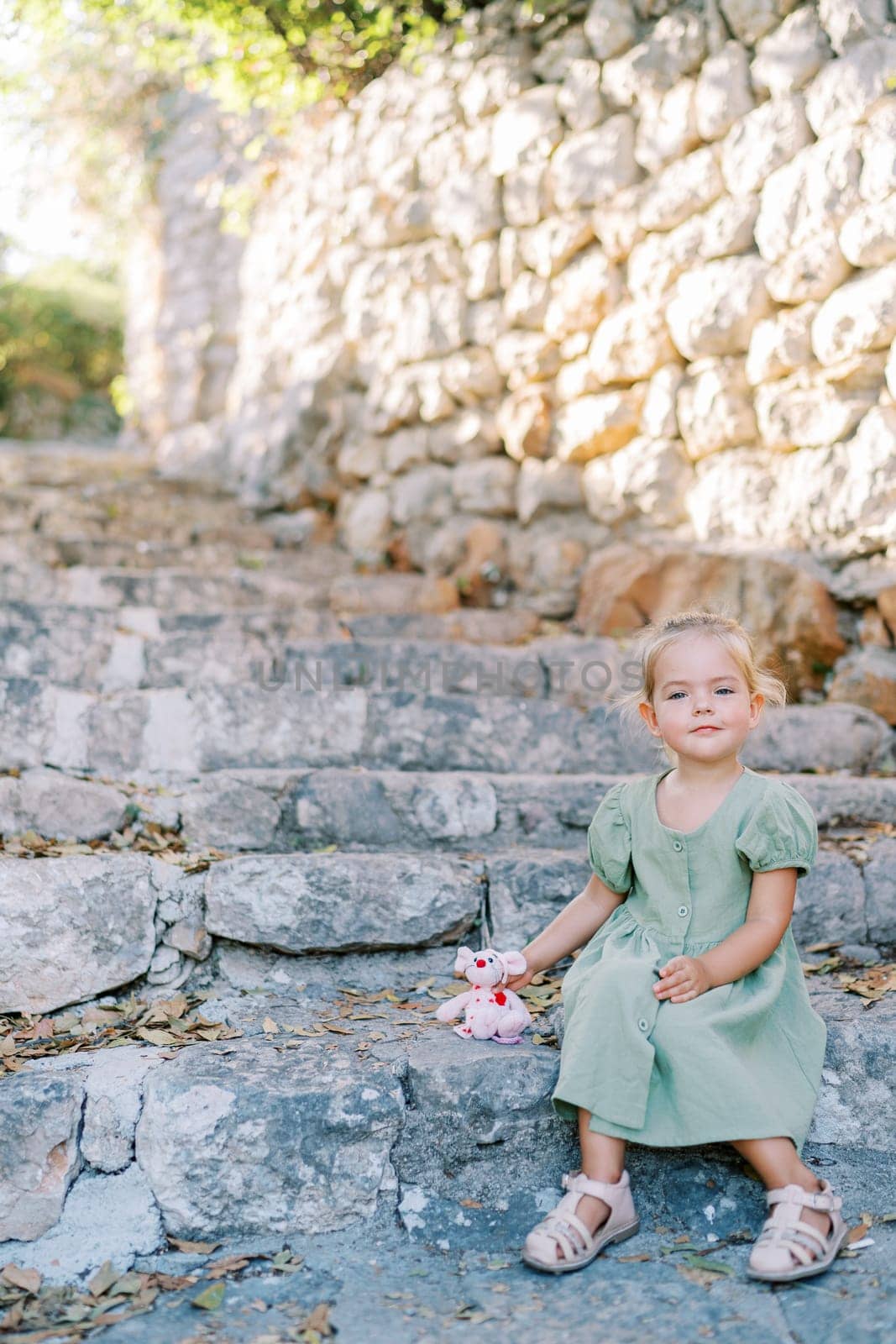 Little smiling girl with toy pink mouse sitting on old stone steps in the garden. High quality photo