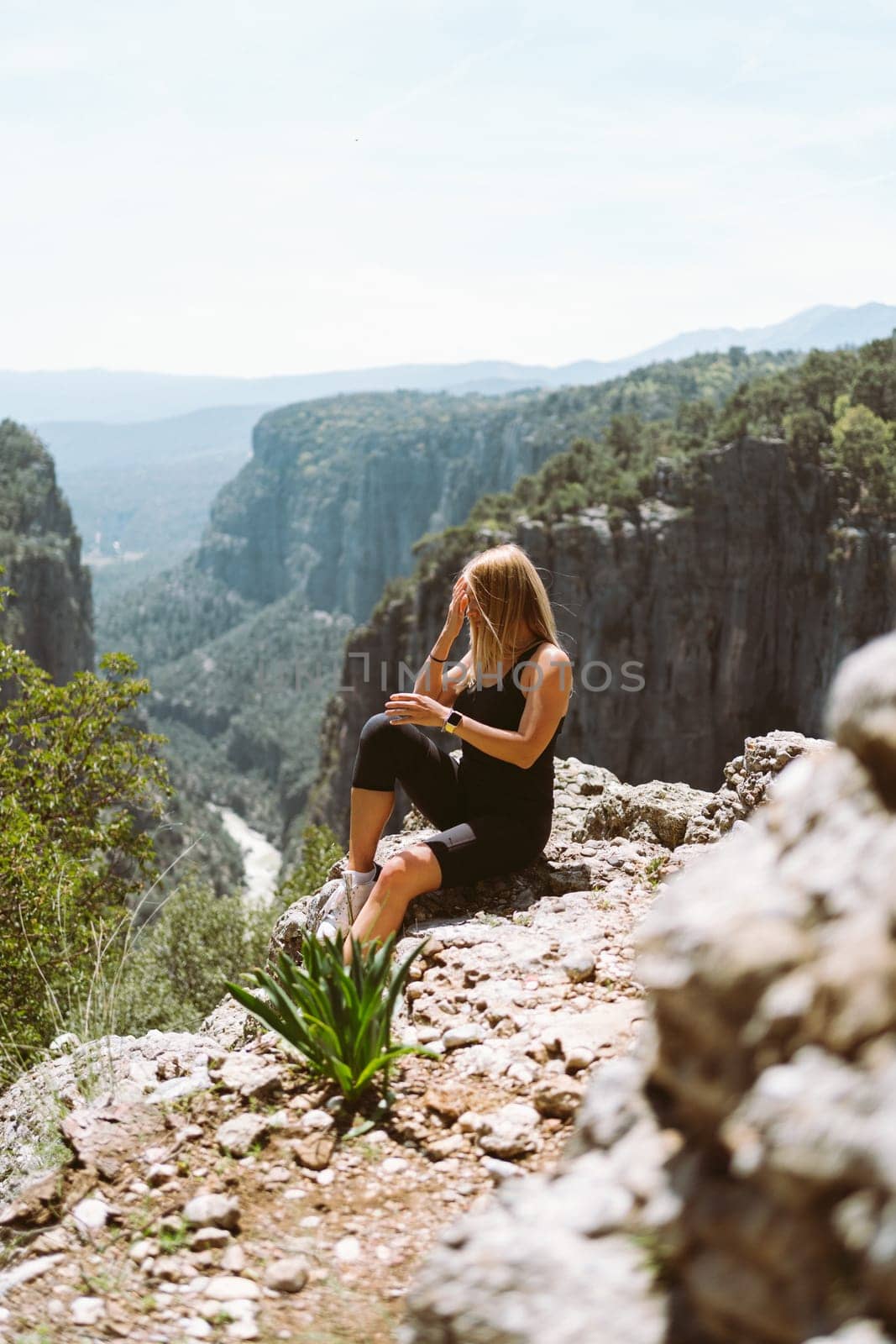 Young beautiful girl woman in black sporty slim outfit sitting on edge rock mountain cliff peak with view on valley canyon. Picture of fit female hiker reached the mountain top by Ostanina