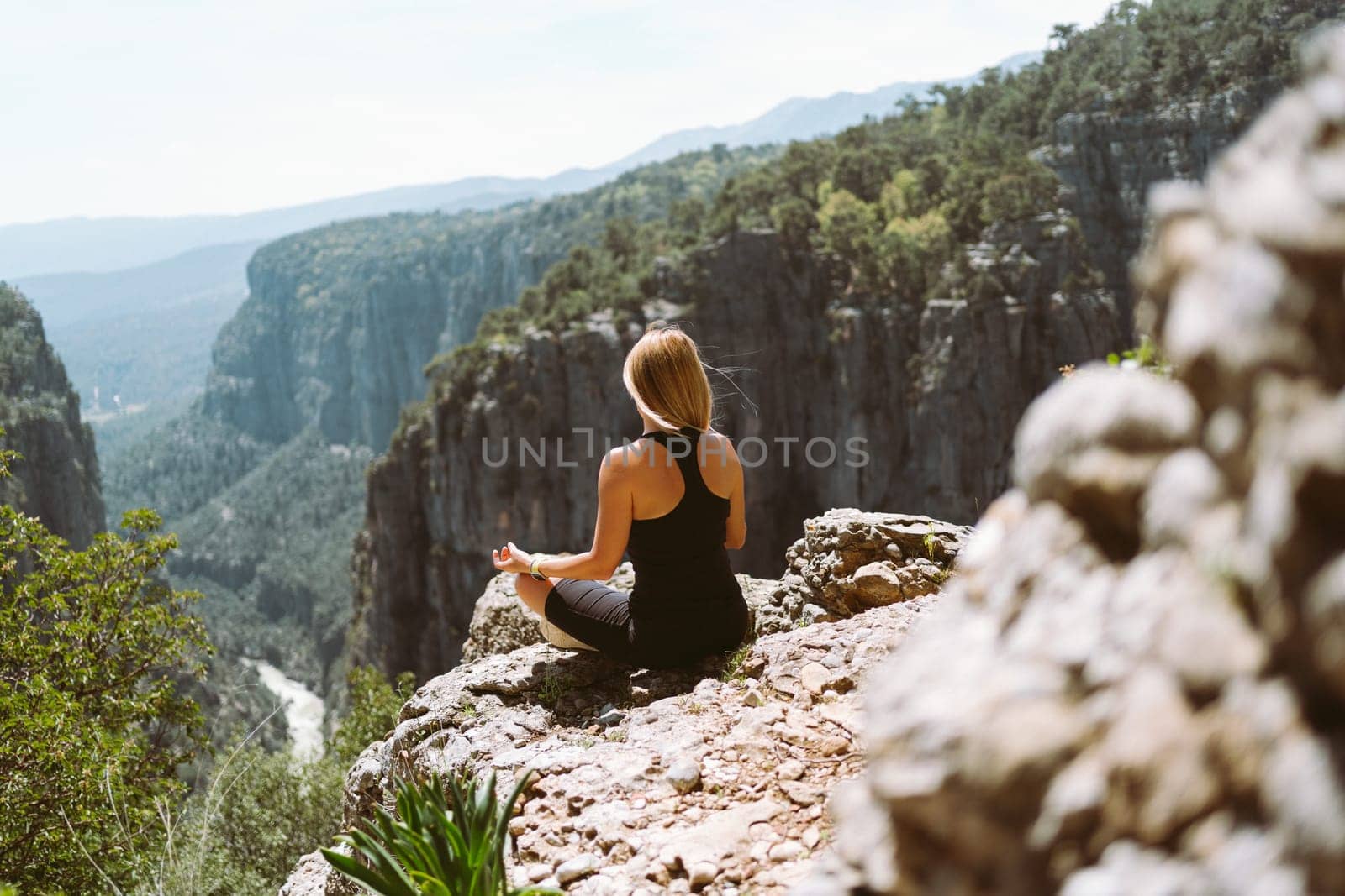 Young beautiful girl woman in black sporty slim outfit meditating on edge rock mountain cliff peak with view on valley canyon. Picture from the back on fit female hiker reached the mountain top by Ostanina